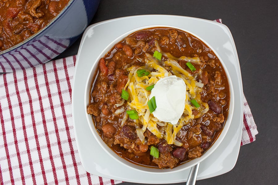 Thick & Hearty Homestyle Chili in a white bowl garnished with sour cream, shredded cheese, and diced green onions
