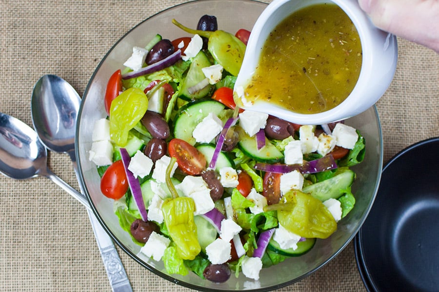 Greek salad in a glass bowl with Greek salad dressing being drizzling over.