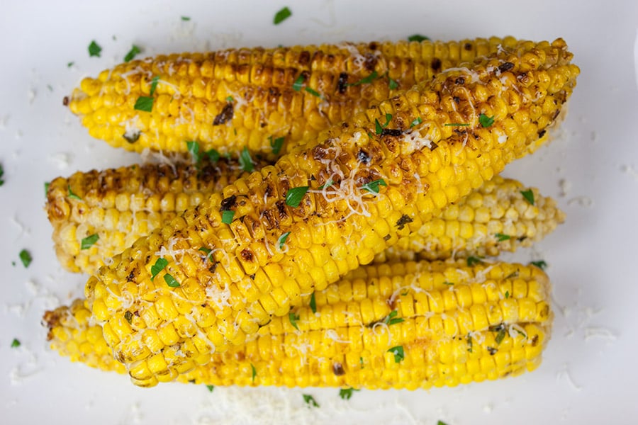 Grilled Parmesan Corn On The Cob on a white platter garnished with parsley and parmesan cheese