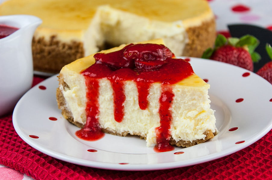 Smooth and Creamy Cheesecake on white plate with red dots drizzled with fresh strawberry coulis