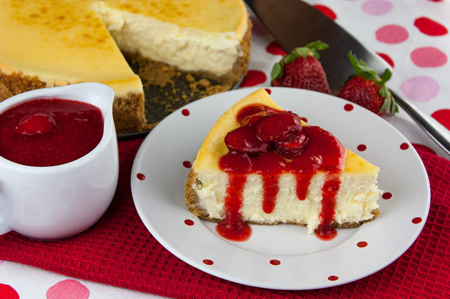 Smooth and Creamy Cheesecake on white plate with red dots drizzled with fresh strawberry coulis