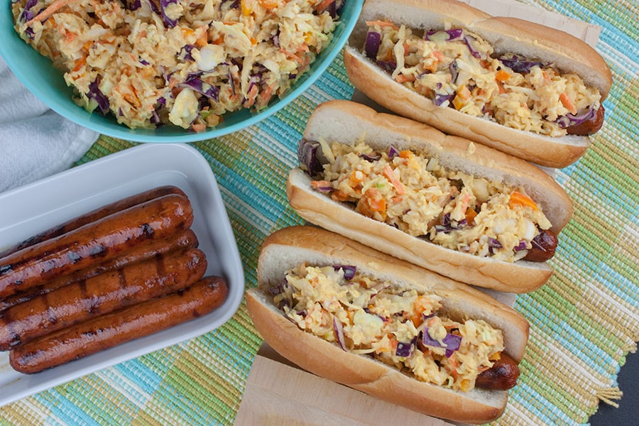Hot Dogs topped with Mustard Slaw on a wooden hot dog board with bowl of mustard slaw in background and platter of grilled hot dogs.