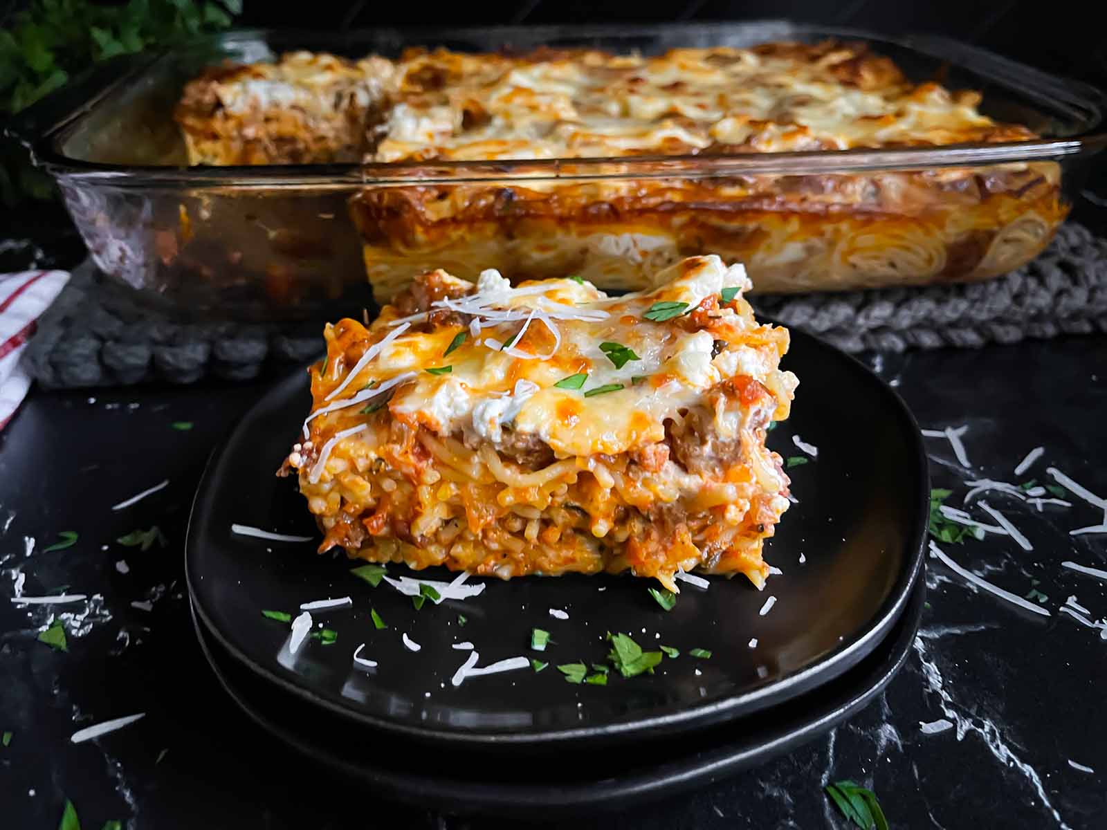 A serving of baked spaghetti on a dark plate garnished with parsley and parmesan cheese.