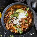 Smoked pork chili garnished with cilantro, onion, lime, sour cream, and onion in a dark bowl.