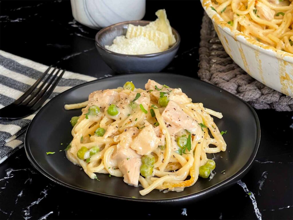 Chicken tetrazzini in a white baking dish with a serving on a dark plate.