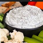 Dill dip in a small dark bowl surrounded by carrots, celery, cauliflower, broccoli, mini bell peppers and rye bread pieces on a dark plate.