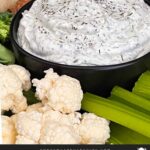 Dill dip in a small dark bowl surrounded by carrots, celery, cauliflower, broccoli, mini bell peppers and rye bread pieces on a dark plate.