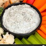 Dill dip in a small dark bowl surrounded by carrots, celery, cauliflower, broccoli, mini bell peppers and rye bread pieces on a dark plate.