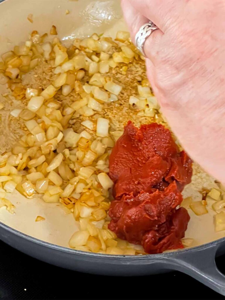 Tomato paste being added to a skillet with onions in it.