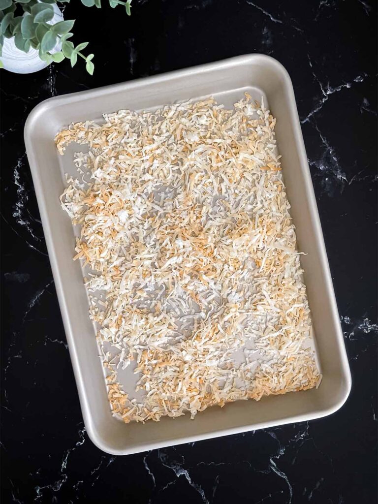 Toasted shredded coconut in a metal baking pan on a dark surface.