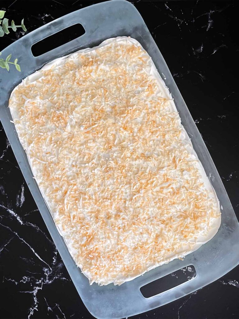 Whipped topping spread over the coconut cream lush and sprinkled with toasted shredded coconut in a glass baking pan on a dark surface.