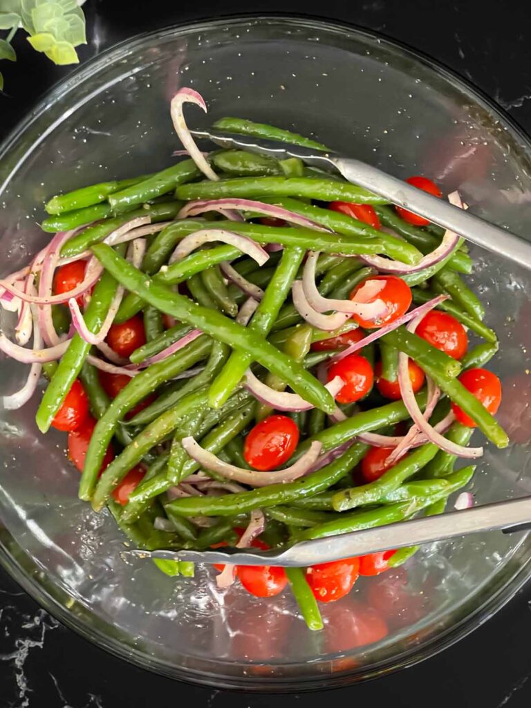 Italian green bean salad tossed with the vinaigrette in a glass mixing bowl on a dark surface.