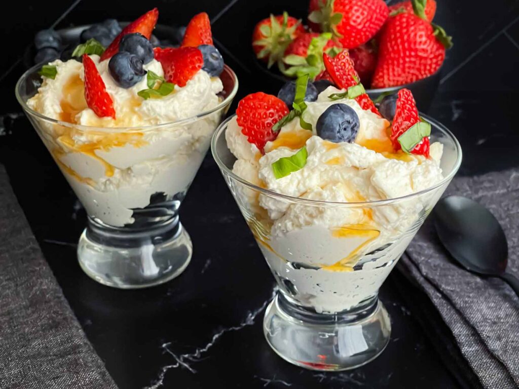 Ricotta mousse in a stemless martini glass garnished with strawberries, blueberries, basil, and honey.