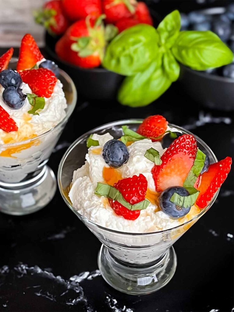 Ricotta mousse in a stemless martini glass garnished with strawberries, blueberries, basil, and honey.