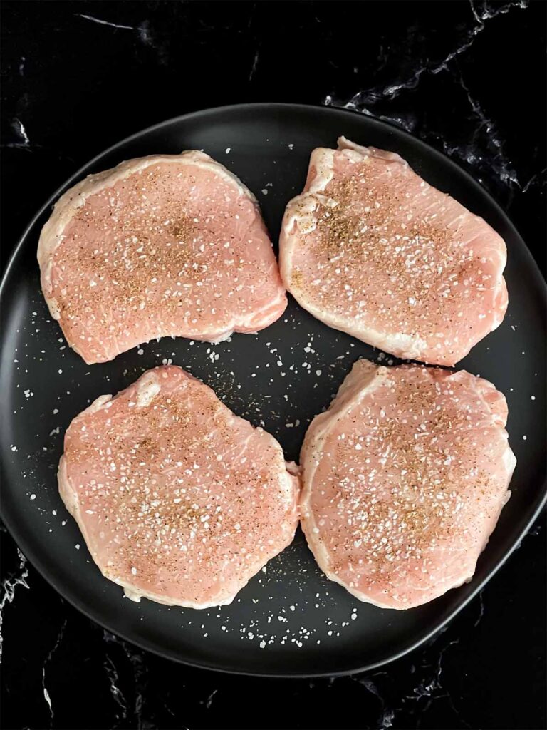 Boneless pork chops seasoned with kosher salt and black pepper on a dark plate.