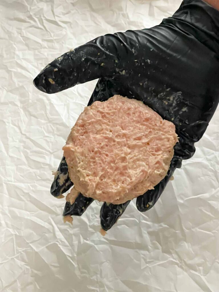 A raw chicken burger patty in a black gloved hand.