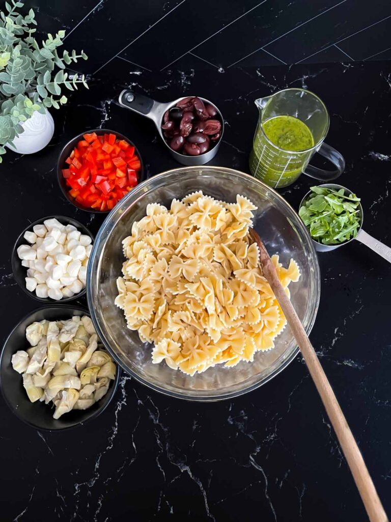 Cooked Farfalle pasta in a glass bowl with the prepped salad ingredients around the bowl.