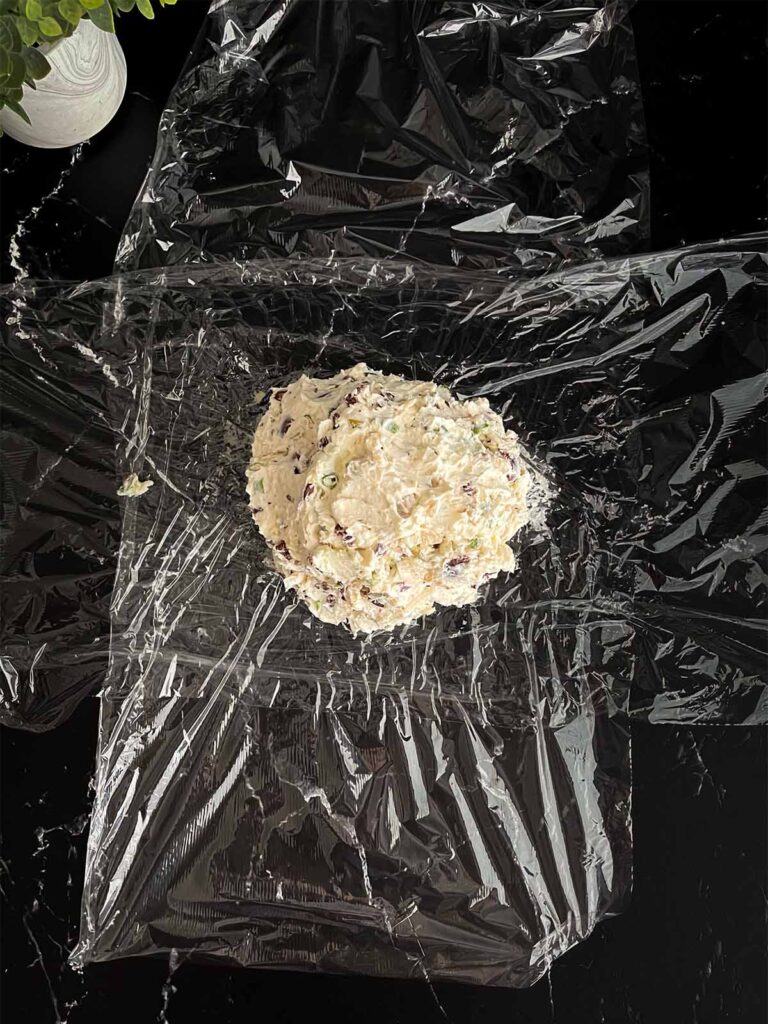 Olive cheese ball mixture placed in the center of two pieces of plastic wrap on a dark surface.