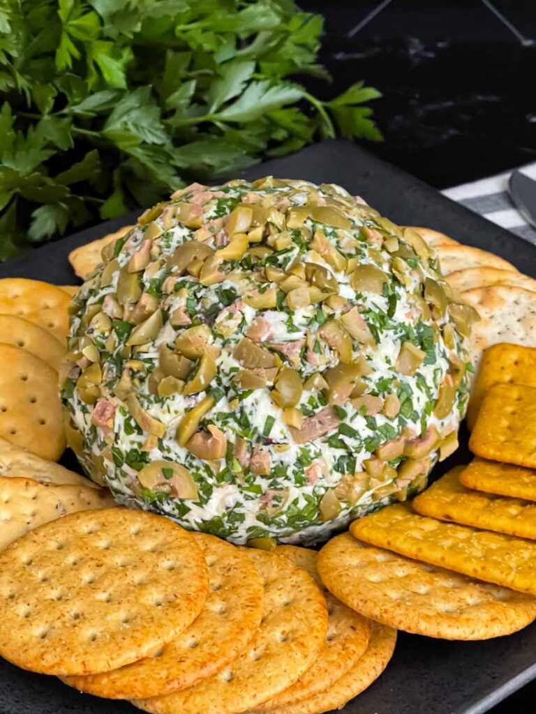 Olive cheese ball on a dark plate surrounded by different shaped crackers.