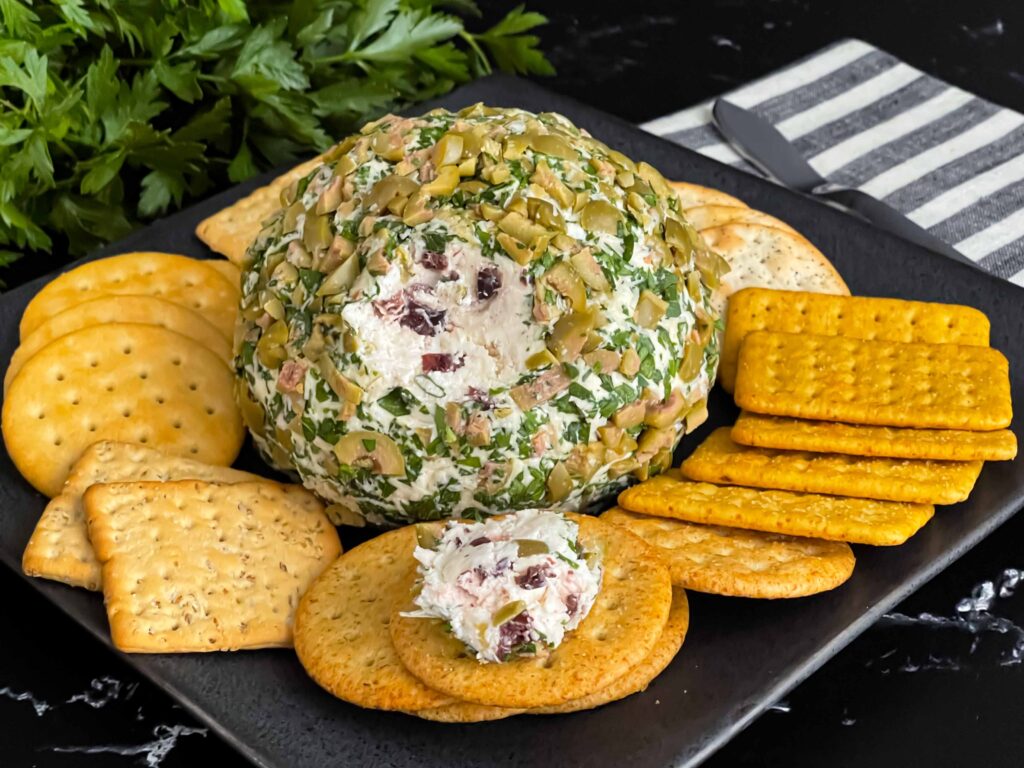 Olive cheese ball on a dark plate surrounded by different shaped crackers.