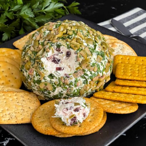 Olive cheese ball on a dark plate surrounded by different shaped crackers.