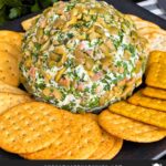 Olive cheese ball on a dark plate surrounded by different shaped crackers.