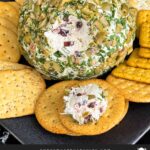 Olive cheese ball on a dark plate surrounded by different shaped crackers.