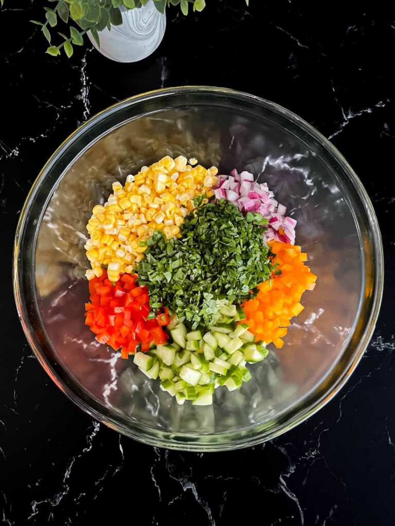 Fresh corn, diced red bell pepper, orange bell pepper, cucumber, red onion, basil, and parsley in a glass mixing bowl.