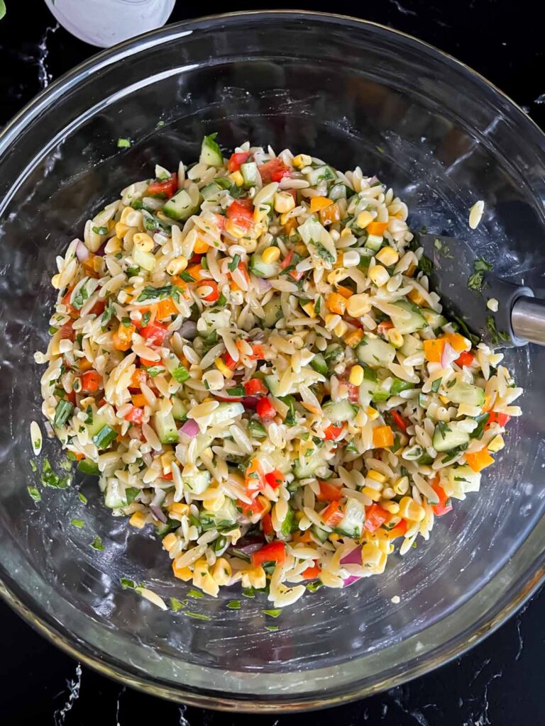 Orzo pasta salad mixed in a glass bowl on a dark surface.