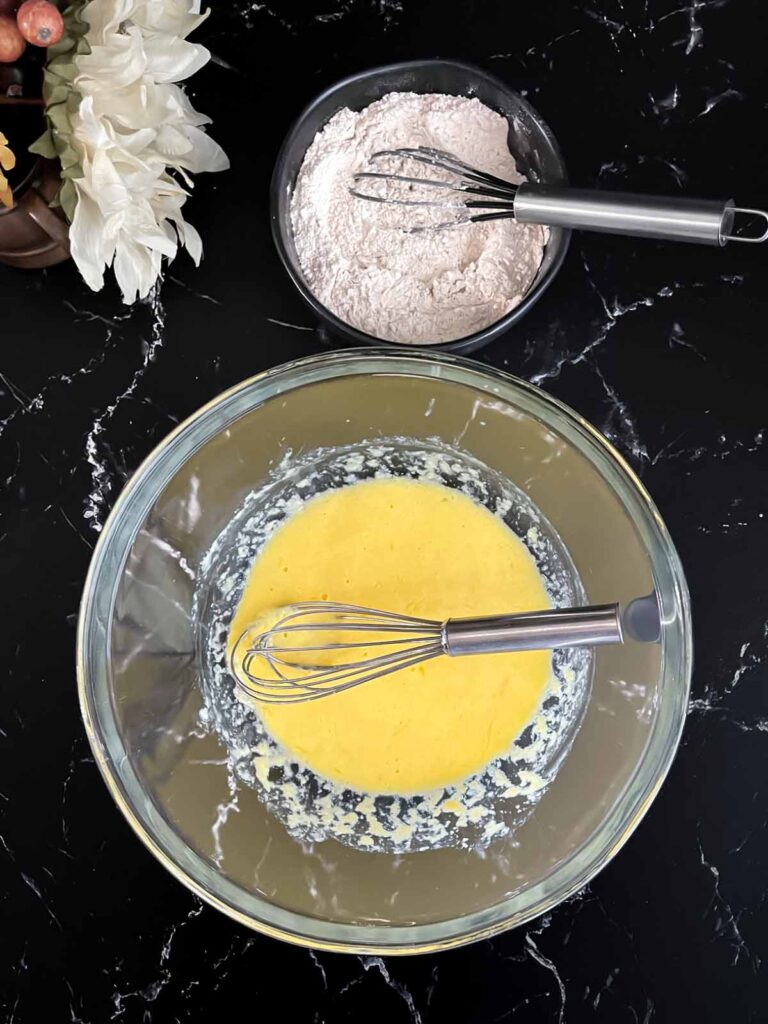 Wet ingredients whisked in a glass bowl and dry ingredients whisked in a dark bowl for the apple fritters recipe.