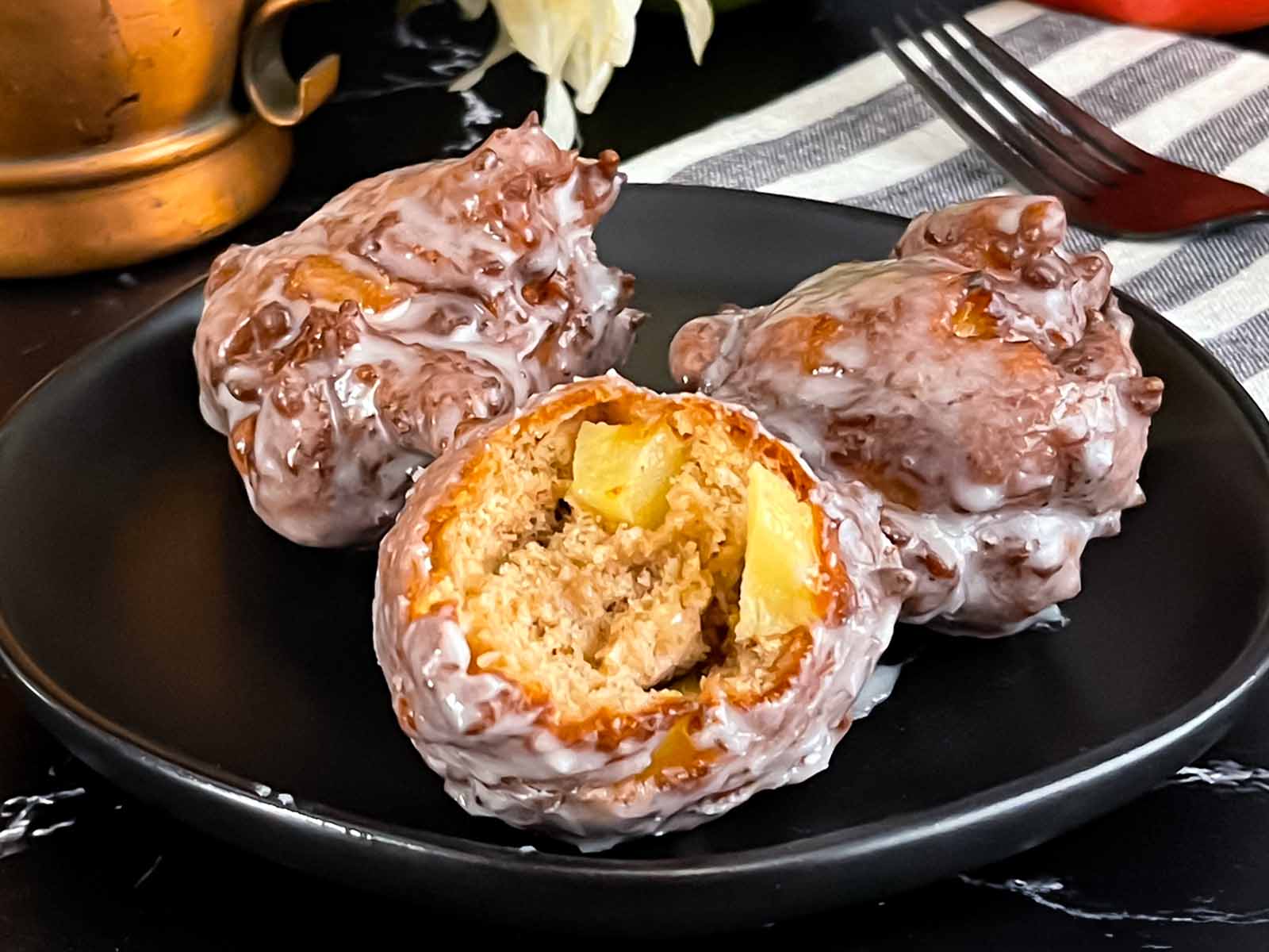 Three apple fritters on a dark plate one with a bite taken.