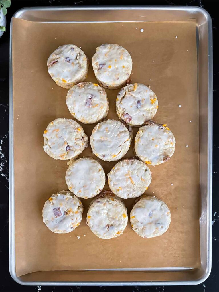 Unbaked bacon cheddar biscuits almost touching on a parchment lined baking sheet.