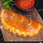 A calzone on a cutting board with a dipping sauce.