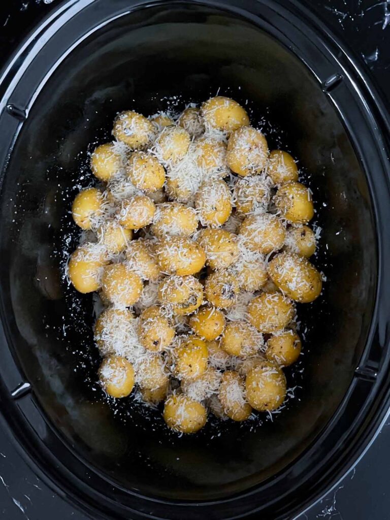 Shredded parmesan sprinkled over the seasoned potatoes in the crock of a slow cooker.