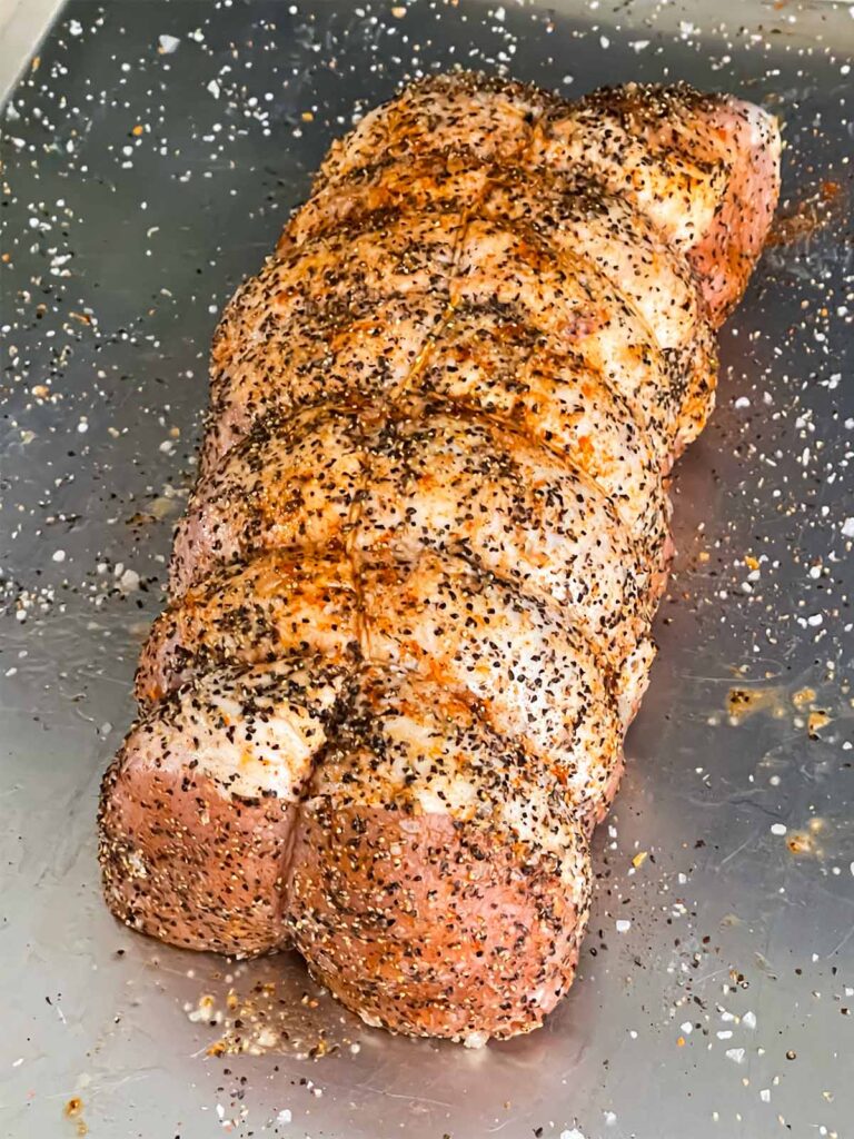 A pork loin trussed and seasoned with salt and black pepper on a baking sheet.