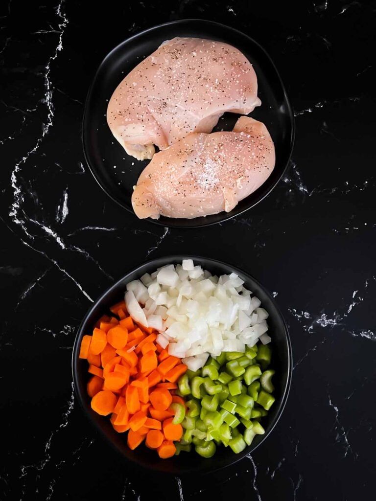 Chicken breasts on a dark plate seasoned with salt and pepper and a dark bowl full of diced onions, carrots, and celery.