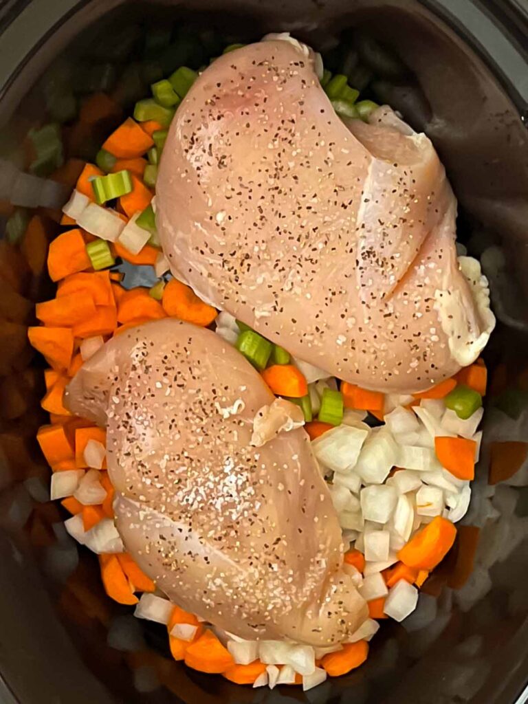 Vegetables in the bottom of a slow cooker with seasoned chicken breasts on top.