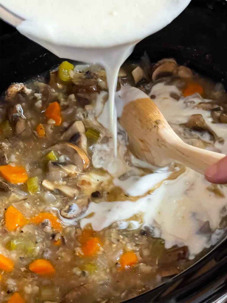 Cream mixture being added to the chicken and wild rice soup in the crock of a slow cooker.