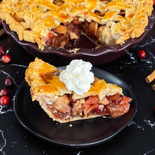 Slice of cranberry apple pie garnished with a dollop of whipped cream on a dark plate.