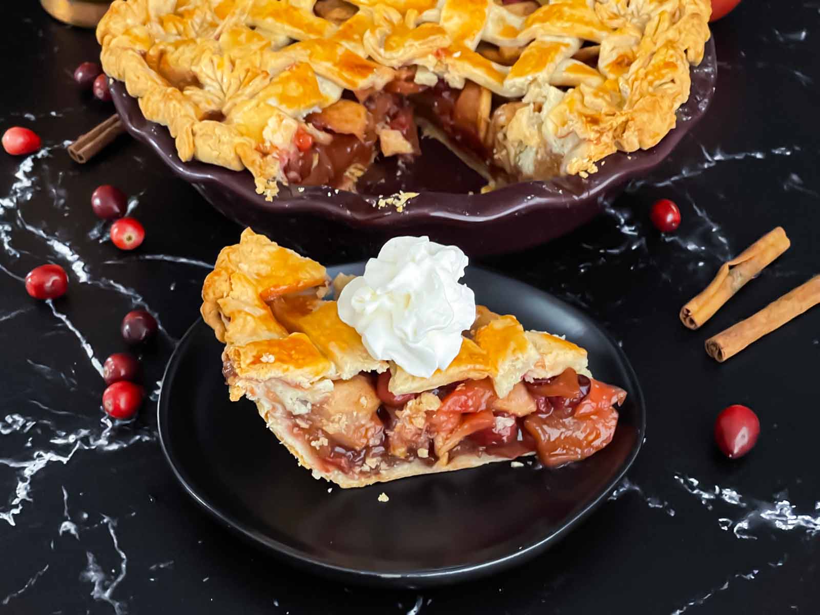 A slice of cranberry apple pie on a dark plate garnished with whipped cream.