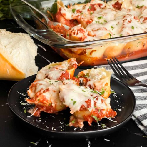 Italian stuffed shells on a black plate with some bread and a casserole dish in the background.