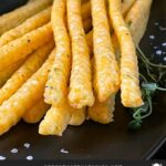 Cheddar and herb cheese straws on a black plate.