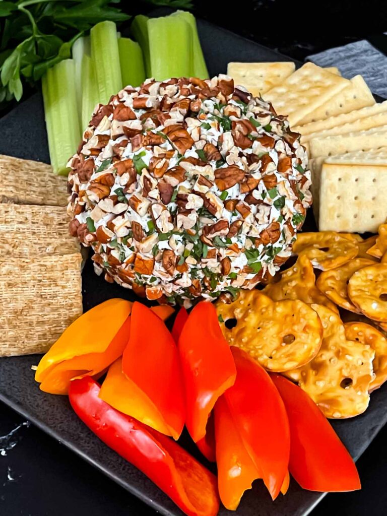 A garlic herb cheese ball on a black plate with crackers and vegetables.