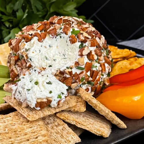 A garlic herb cheese ball on a black plate with crackers and vegetables.