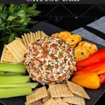 A garlic herb cheese ball on a black plate with crackers and vegetables.