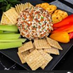 A garlic herb cheese ball on a black plate with crackers and vegetables.