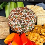 A garlic herb cheese ball on a black plate with crackers and vegetables.