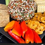 A garlic herb cheese ball on a black plate with crackers and vegetables.